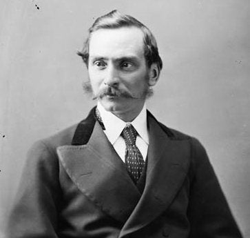 A vintage black and white portrait photograph of a man with a mustache, wearing a dark suit jacket, white shirt, and patterned tie against a plain studio background.
